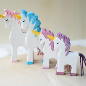 May include: Four wooden unicorn figurines in a row. The unicorns are white with colorful manes and tails. The manes are pink, blue, and purple. The horns are yellow, and the hooves are pink and blue.