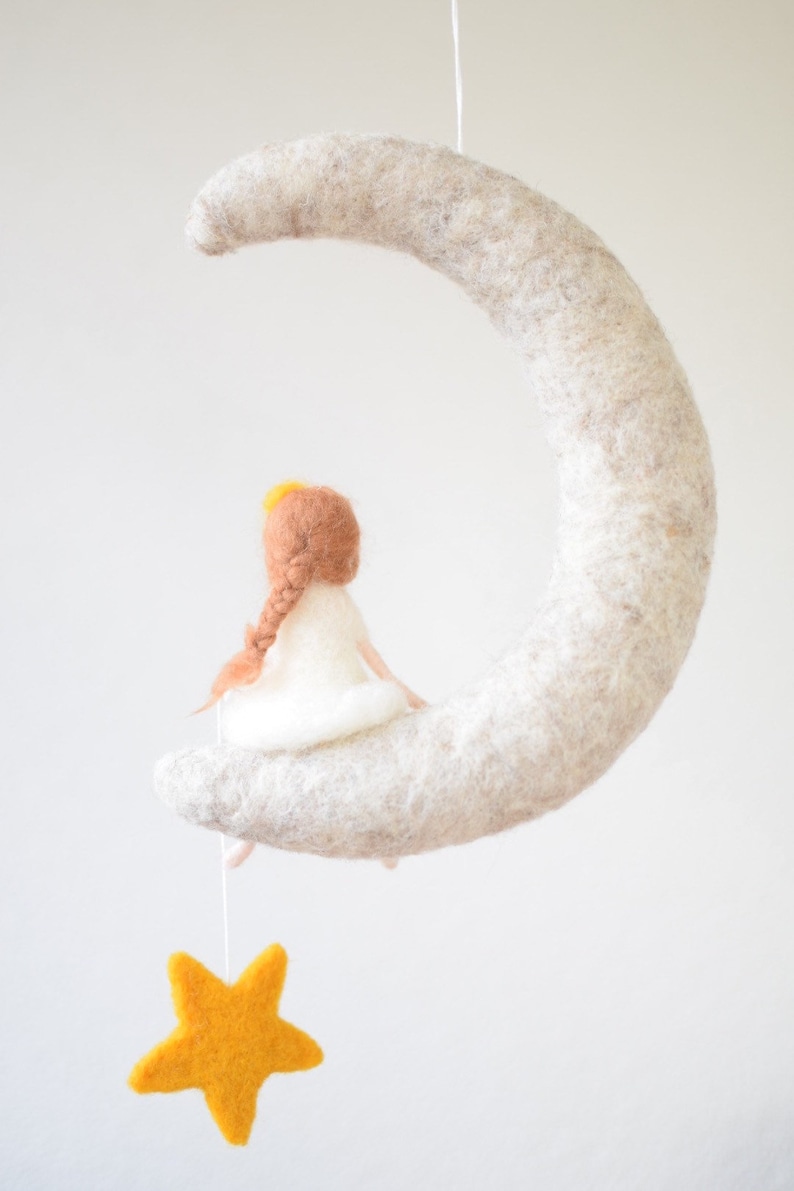 May include: A felt moon mobile with a small felt girl sitting on it. The girl has brown hair and a white dress. A yellow felt star hangs from the moon.