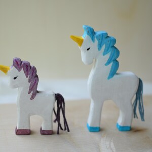 May include: Two wooden unicorn figurines. One has a purple mane and tail, the other has a blue mane and tail. Both have yellow horns, white bodies, and are painted with pink and blue accents. They stand on a light-colored wooden surface.