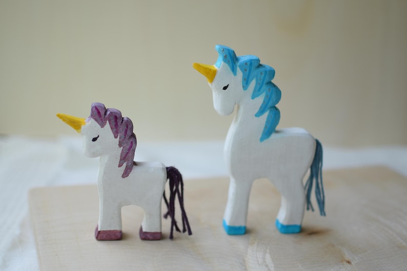 May include: Two wooden unicorn figurines. One has a purple mane and tail, the other has a blue mane and tail. Both have yellow horns, white bodies, and are painted with pink and blue accents. They stand on a light-colored wooden surface.
