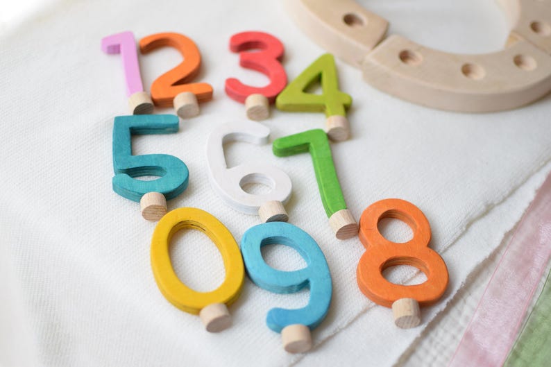 Pu&ograve; includere: Una collezione di dieci numeri in legno da zero a nove, ognuno dipinto in un colore distinto tra cui rosa, arancione, rosso, verde lime, verde acqua, bianco, giallo e blu. Ogni numero presenta un piccolo piolo cilindrico in legno alla base.