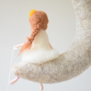 May include: A felt doll with brown hair and a white dress sits on a crescent moon shaped felt object. The doll is holding a string in its hand.