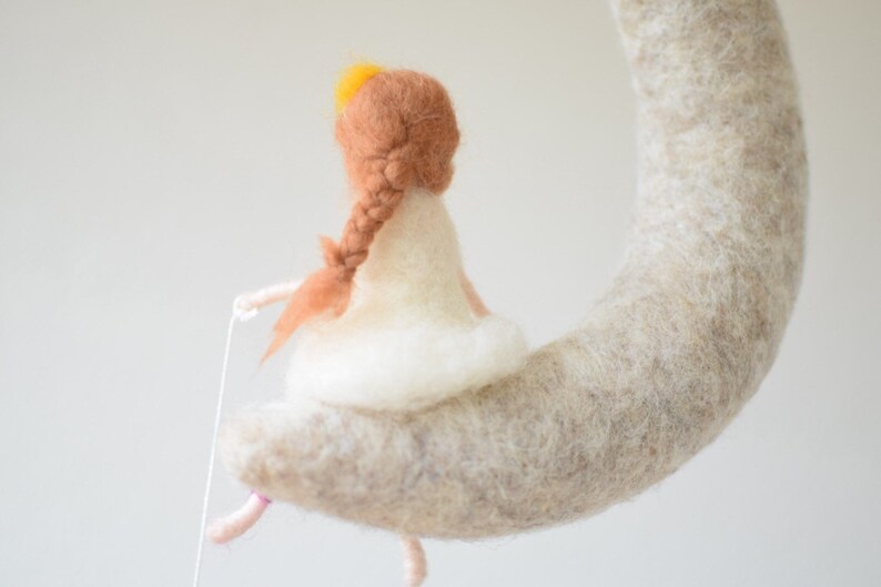 May include: A felt doll with brown hair and a white dress sits on a crescent moon shaped felt object. The doll is holding a string in its hand.