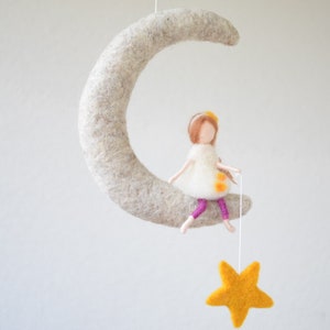 May include: A felt doll with brown hair and a white dress sits on a felt crescent moon. The doll is holding a string attached to a yellow felt star.