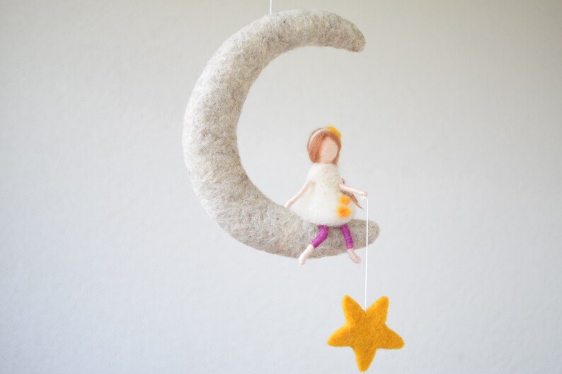 May include: A felt doll with brown hair and a white dress sits on a felt crescent moon. The doll is holding a string attached to a yellow felt star.