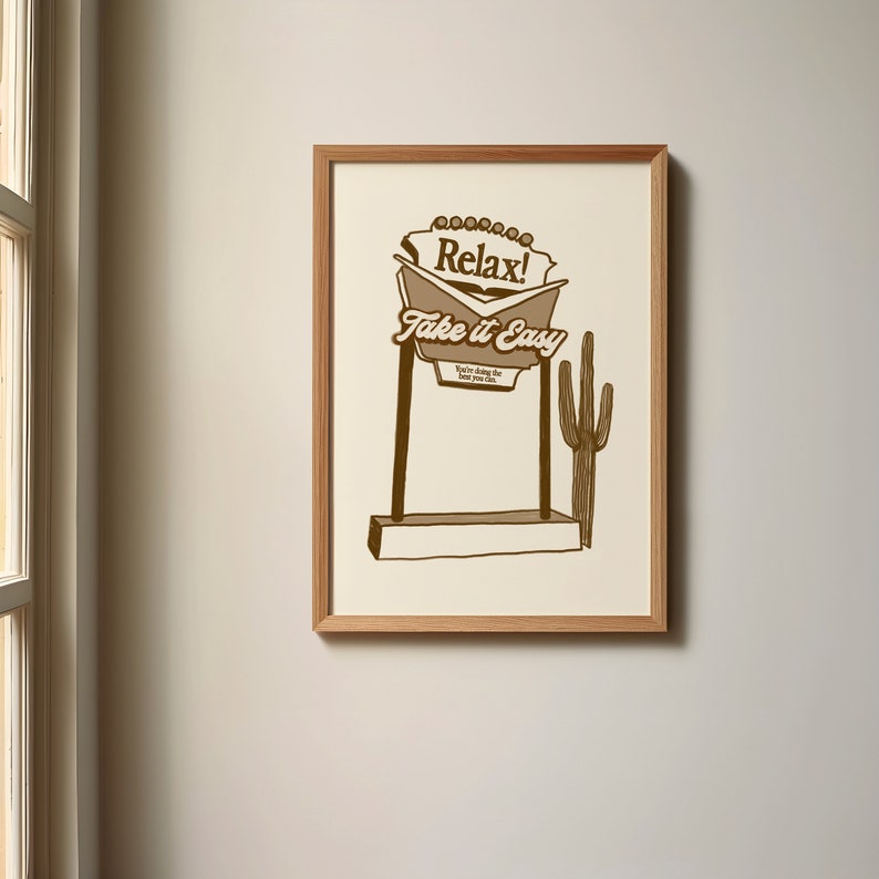 Op de afbeelding: Een ingelijst print met een bruine illustratie van een woestijnwegwijzer met de tekst "Relax! Take it Easy. You're doing the best you can." Het bord staat voor een bruine cactus.