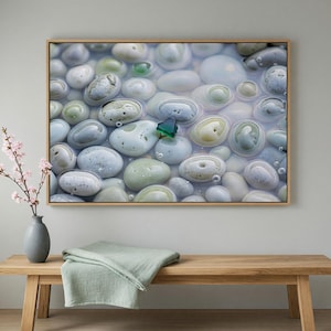 May include: Framed artwork featuring a close-up of smooth, water-covered stones in shades of gray, green, and white. A small, green, triangular object is visible among the stones. The artwork is displayed above a wooden bench with a folded green blanket.
