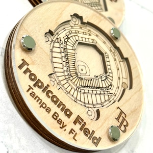 May include: A wooden coaster with a laser-engraved design of Tropicana Field, home of the Tampa Bay Rays baseball team. The coaster features the team's logo, "TB", and the stadium's name and location.