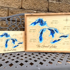 May include: Two wooden maps of the Great Lakes with blue water features. The larger map is framed in wood and the smaller map is unframed. Both maps have the text "The Great Lakes" printed on them.