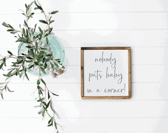 May include: A square wooden sign with a dark brown frame and a white background featuring the black cursive text 'nobody puts baby in a corner!' centered on the surface. The sign is displayed next to a glass vase containing green olive branches.