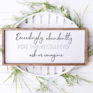 May include: A wooden sign with the text "Exceedingly, abundantly more than we could ever ask or imagine" in black script font. The sign is surrounded by white flowers and greenery.