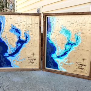 May include: Two rectangular wooden wall maps of Tampa Bay, Florida, featuring layered laser-cut wood with varying shades of blue to represent water depths. Each map includes engraved street names, a compass rose, and the text Tampa Bay.