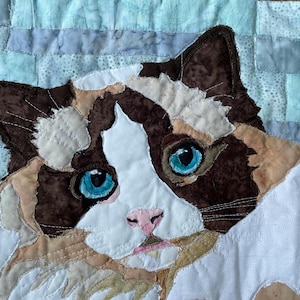 May include: A close-up of a quilted cat's face, with brown and white fur, bright blue eyes, and a pink nose. The background is a patchwork of light blue and white fabric squares. The artwork is a textile portrait of a cat.