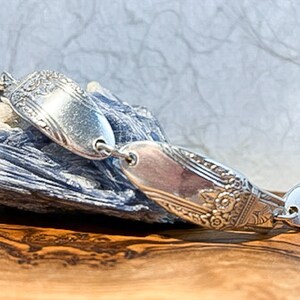May include: A silver bracelet made from repurposed silverware. The bracelet is made of three spoon bowls, each with a different design. The bracelet is on a blue and gray rock.