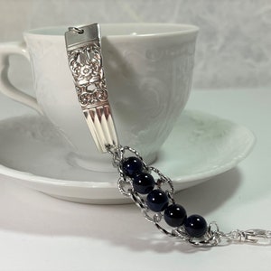 May include: A silver bracelet with a decorative spoon charm and a chain of dark blue beads. The bracelet has a silver clasp.