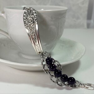 May include: A silver spoon bracelet with a floral design and black beads. The bracelet is on a white saucer.