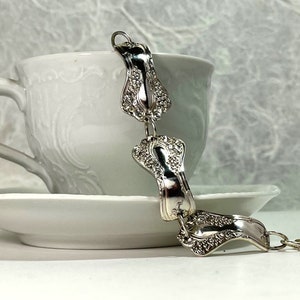 May include: A silver bracelet made from repurposed silverware. The bracelet is made of several links, each with a unique design. The bracelet is resting on a white teacup and saucer.