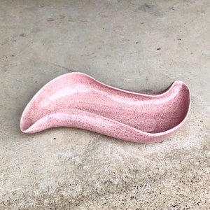 Puede incluir: Un plato de cerámica rosa de estilo moderno de mediados de siglo con un acabado moteado. El plato tiene una forma curva única con bordes elevados. El color es un rosa suave y polvoriento con motas oscuras.