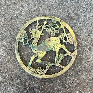 May include: A circular, brass-toned decorative item depicting a deer leaping amidst floral and leaf motifs. The deer is centrally positioned within a circular frame. The piece has a weathered, antique appearance, with a verdigris hue.