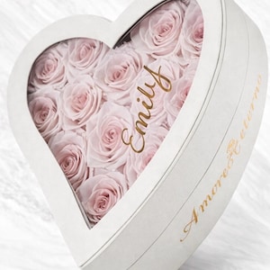 May include: A white heart-shaped box filled with pale pink roses. The box has a clear window, and the name "Emily" is written in gold script. The words "Amore Eterno" are also written in gold script on the side of the box.