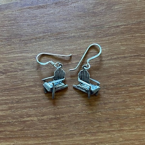 May include: A pair of silver-colored Adirondack chair earrings. The earrings have a detailed design, with the chair's back, seat, and armrests clearly visible. The earrings are suspended from a simple hook.