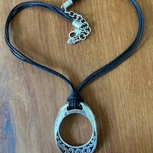 May include: A silver-toned pendant necklace with a black cord. The pendant is an oval shape with a decorative scroll design. The necklace cord is arranged in a heart shape.