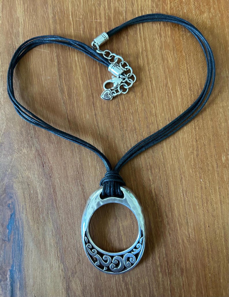 May include: A silver-toned pendant necklace with a black cord. The pendant is an oval shape with a decorative scroll design. The necklace cord is arranged in a heart shape.