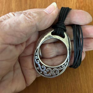 May include: A silver-toned pendant necklace with a large, oval-shaped pendant. The pendant features a decorative, openwork design with swirling patterns. The necklace has a black cord. The pendant is held in a hand.