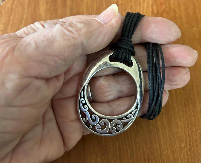 May include: A silver-toned pendant necklace with a large, oval-shaped pendant. The pendant features a decorative, openwork design with swirling patterns. The necklace has a black cord. The pendant is held in a hand.
