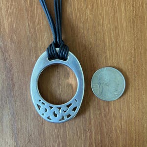 May include: Silver-toned oval pendant necklace with a decorative cutout design on the lower half. The pendant is suspended from a black cord. A U.S. quarter coin is next to the pendant for size comparison.