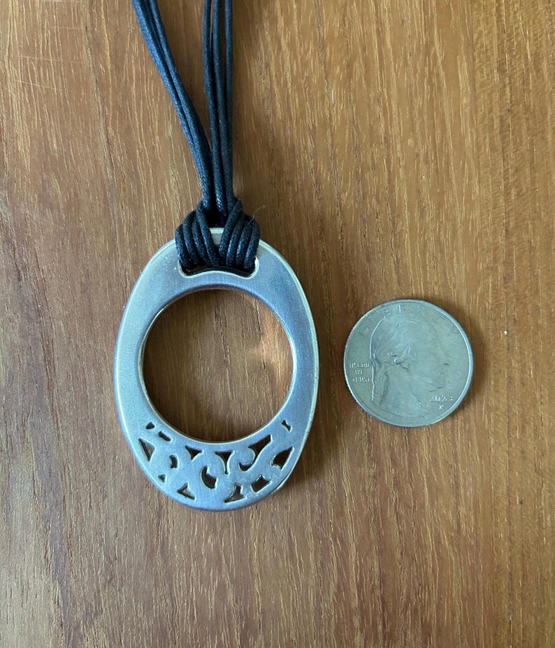 May include: Silver-toned oval pendant necklace with a decorative cutout design on the lower half. The pendant is suspended from a black cord. A U.S. quarter coin is next to the pendant for size comparison.