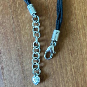 May include: Close-up of a black cord necklace with silver-toned metal accents. The necklace features a chain extender with a heart-shaped charm and a lobster clasp closure. The cord is braided and appears to be made of leather or a similar material.