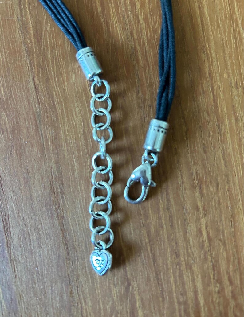 May include: Close-up of a black cord necklace with silver-toned metal accents. The necklace features a chain extender with a heart-shaped charm and a lobster clasp closure. The cord is braided and appears to be made of leather or a similar material.