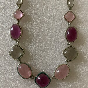 May include: A silver necklace with a variety of colored glass stones. The stones are in shades of pink, purple, and gray. The necklace is made of a silver chain with a clasp.