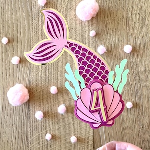 Op de afbeelding: Een roze en paarse zeemeerminstaart taarttopper met een gouden omtrek, een zeeschelp en het nummer 4. De topper staat op een doorzichtige stok, met roze pompons verspreid op een houten oppervlak. Thema: verjaardag.