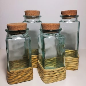 May include: Four clear glass jars with cork lids and a woven straw base. The jars are empty and have a rustic look.