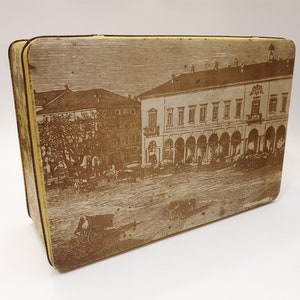 May include: A vintage tin box with a brown and white image of a European city square. The image shows a building with arched doorways and windows, and people walking in the street.