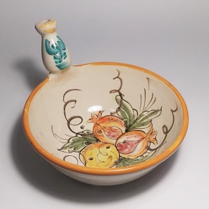 May include: A white ceramic bowl with a handle, decorated with a floral design featuring yellow, orange, and green pomegranates. The bowl has a yellow rim and a white background.