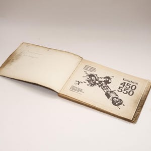 K&ouml;nnte beinhalten: Ein ge&ouml;ffnetes Buch mit einer Schwarzwei&szlig;illustration eines Motorradmotors. Der Buchumschlag tr&auml;gt den Text "Lambro 450 550" in einer fetten, serifenlosen Schrift.