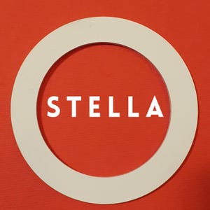 Könnte beinhalten: Ein weißer Kreisring mit einem großen inneren Kreis. Das Wort "STELLA" ist in weißen Buchstaben auf rotem Hintergrund innerhalb des inneren Kreises gedruckt.