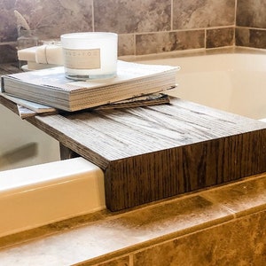 Puede incluir: Una bandeja de baño de madera marrón oscuro, con una pila de revistas, una vela blanca y jabón. La bandeja descansa sobre el borde de una bañera blanca, creando un ambiente de spa.