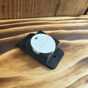 Könnte beinhalten: Eine schwarze Wandhalterung aus Kunststoff mit einem weißen, runden Chippolo-Tracker. Der Tracker trägt die Aufschrift "chippolo".