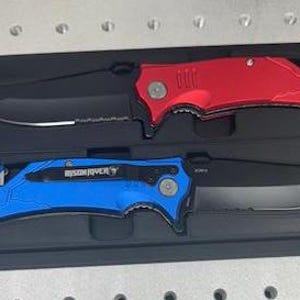 May include: Two pocket knives with black blades and handles in different colors. The top knife has a red handle and the bottom knife has a blue handle. Both knives have a safety feature and a serrated edge.
