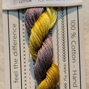Pode incluir: Meada de linha de bordar de algodão torcido em tons de ouro e roxo, rotulada como "VD 13 Oriole". O rótulo inclui texto sobre o algodão tingido à mão na Austrália e comprimentos pré-cortados de 50 cm.
