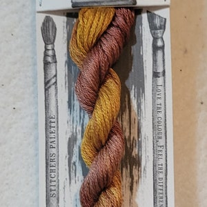 May include: A skein of hand-dyed embroidery floss in caramel and gold tones. The label reads "CARAMEL FUDGE" and "HAND DYED". The packaging features illustrations of paintbrushes and the text "STITCHERS PALETTE".