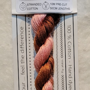 Pode incluir: Um close-up de um novelo de fio de bordado tingido à mão em tons de rosa e marrom. O fio é etiquetado como "VD 07 Hakea Pincushion" e é 100% algodão. Tem 10 metros de comprimento e vem em comprimentos de 50 cm.