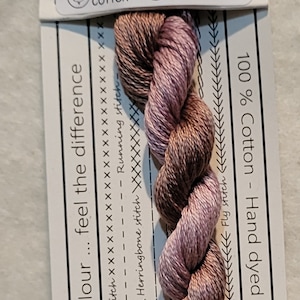 Pode incluir: Meada de linha de bordar de algodão torcido em tons de castanho e malva, rotulada como "VD 21 HOYA". O rótulo inclui texto sobre o algodão e vários pontos de bordado. A linha tem 50 cm de comprimento.