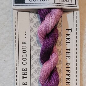May include: A skein of hand-dyed stranded cotton embroidery floss in shades of purple and pink. The floss is labeled "Dahlia" and is 10 meters long. The label also says "Love the Colour... Feel the Difference" and "CGT Hand Dyed".