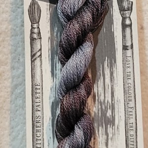 May include: A skein of hand-dyed embroidery floss in shades of purple and blue. The floss is labeled "Storm" and is part of the "Stitchers Palette" collection.