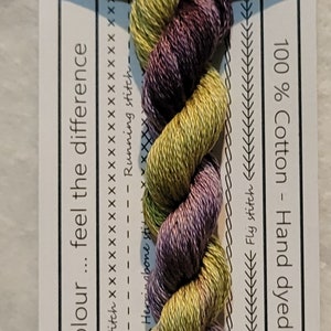 Pode incluir: Um novelo de linha de bordar de algodão torcido em tons de verde e roxo, rotulado como "VD 20 Wompoo Dove". A etiqueta indica 10 m de comprimentos pré-cortados de 50 cm. A linha é 100% algodão e tingida à mão na Austrália. A embalagem inclui exemplos de pontos.